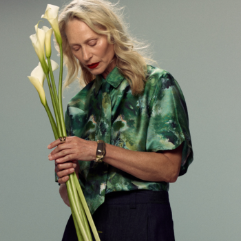 An older woman in a green floral blouse holds a bunch of white calla lilies against a plain grey background.